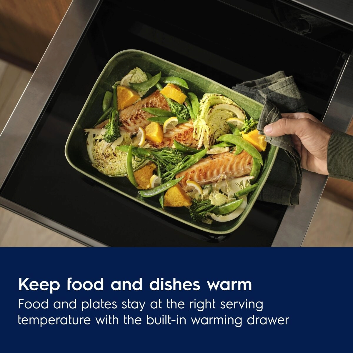Person holding a green dish with food in an oven, with text about Electrolux 600 Series Built In Warming Drawer