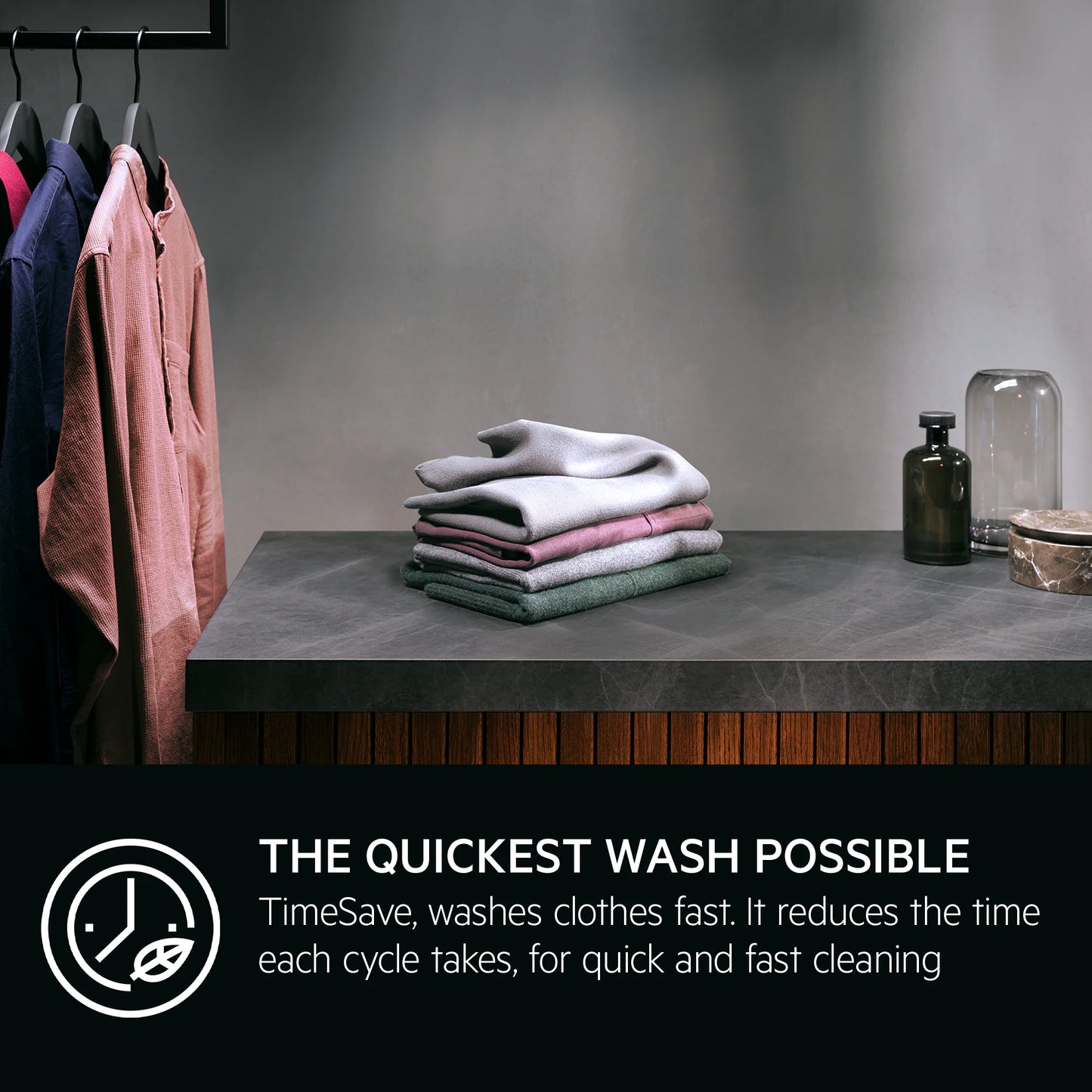 Stack of folded clothes on a table with hanging clothes and decor items in the background. Regarding AEG Quick Wash.